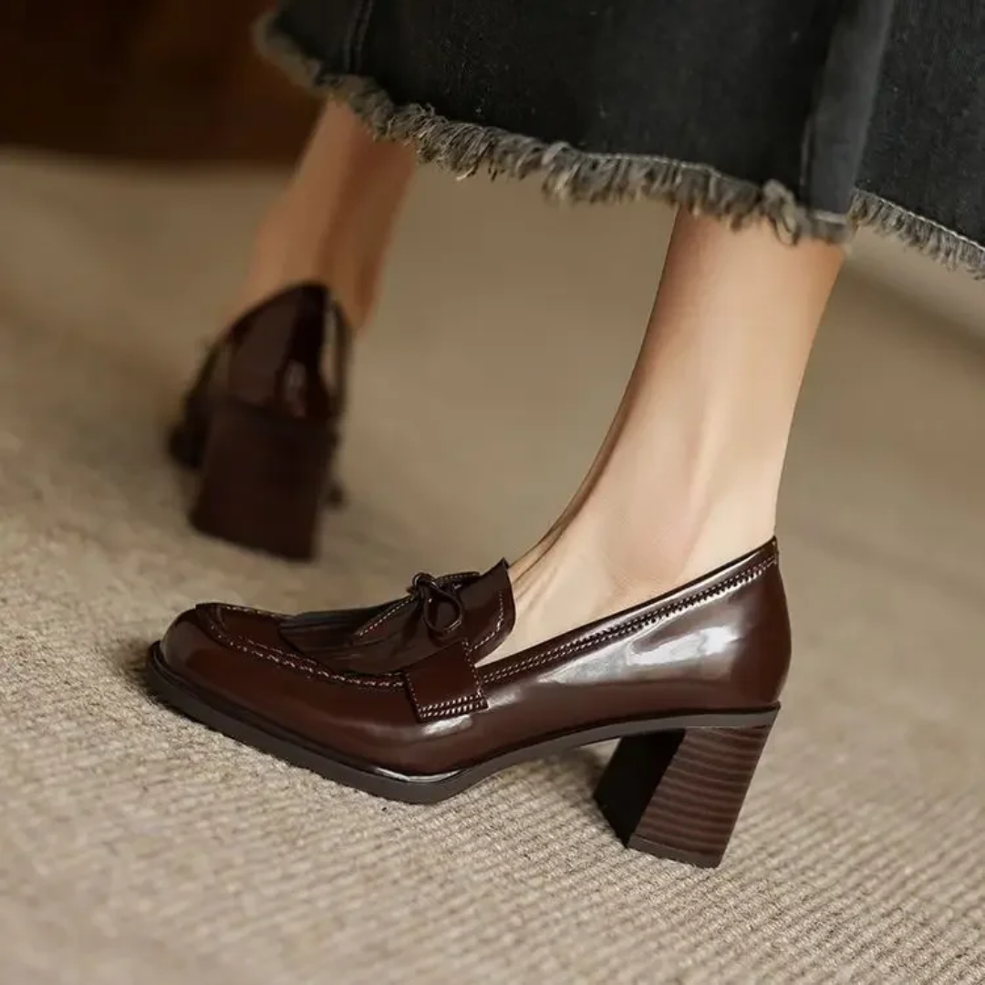 Female model wearing dark block heel loafers with tassel trim on a plain background.