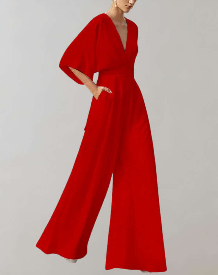 Female model in a flowing wide leg jumpsuit with a deep neckline on a plain background.