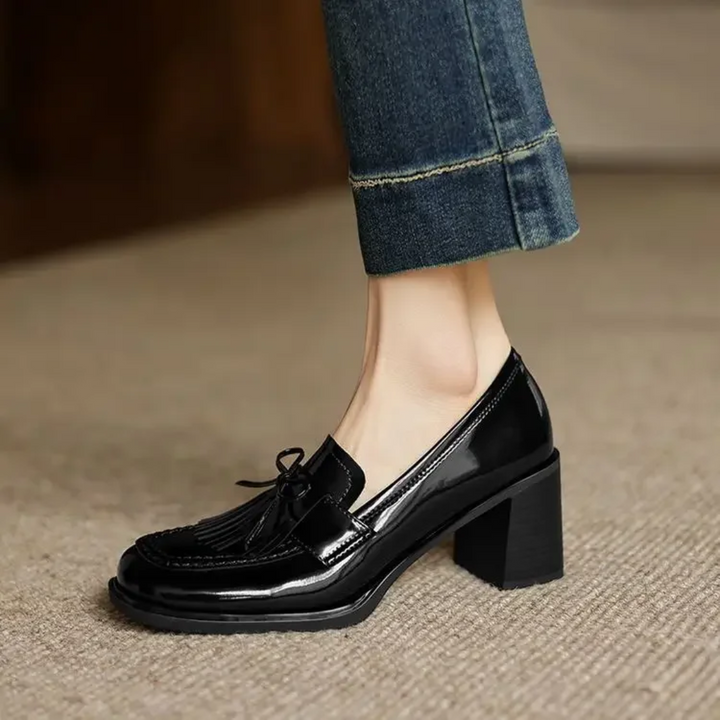 Female modelling dark tassel loafer heels with a round toe on a plain background.