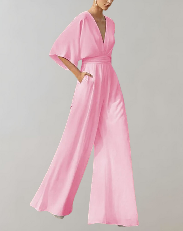 Woman in a deep V neck jumpsuit with wide legs and soft draped sleeves on a plain background.