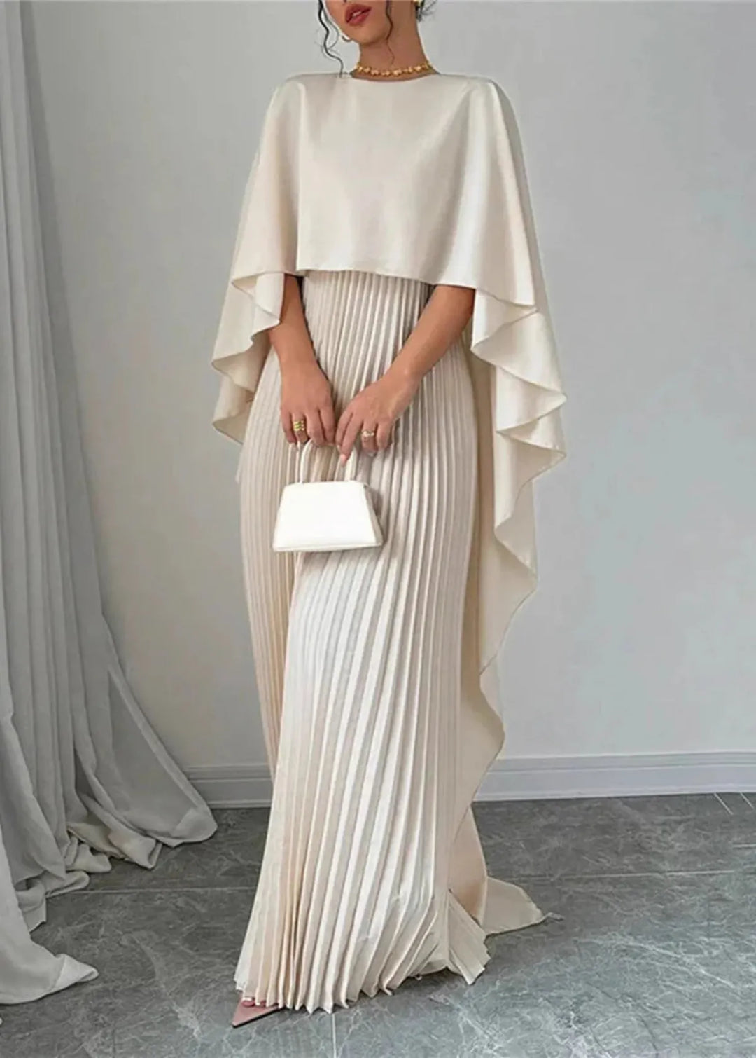 Woman showcasing a cape overlay dress with flowing pleats on a plain background.