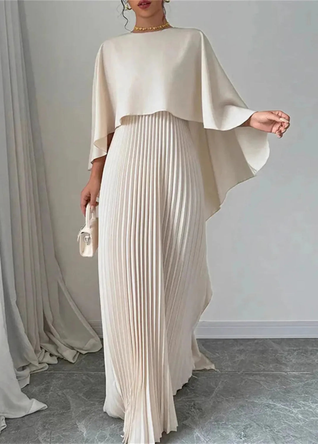 Woman wearing a long pleated dress with cape-style overlay on a plain background.