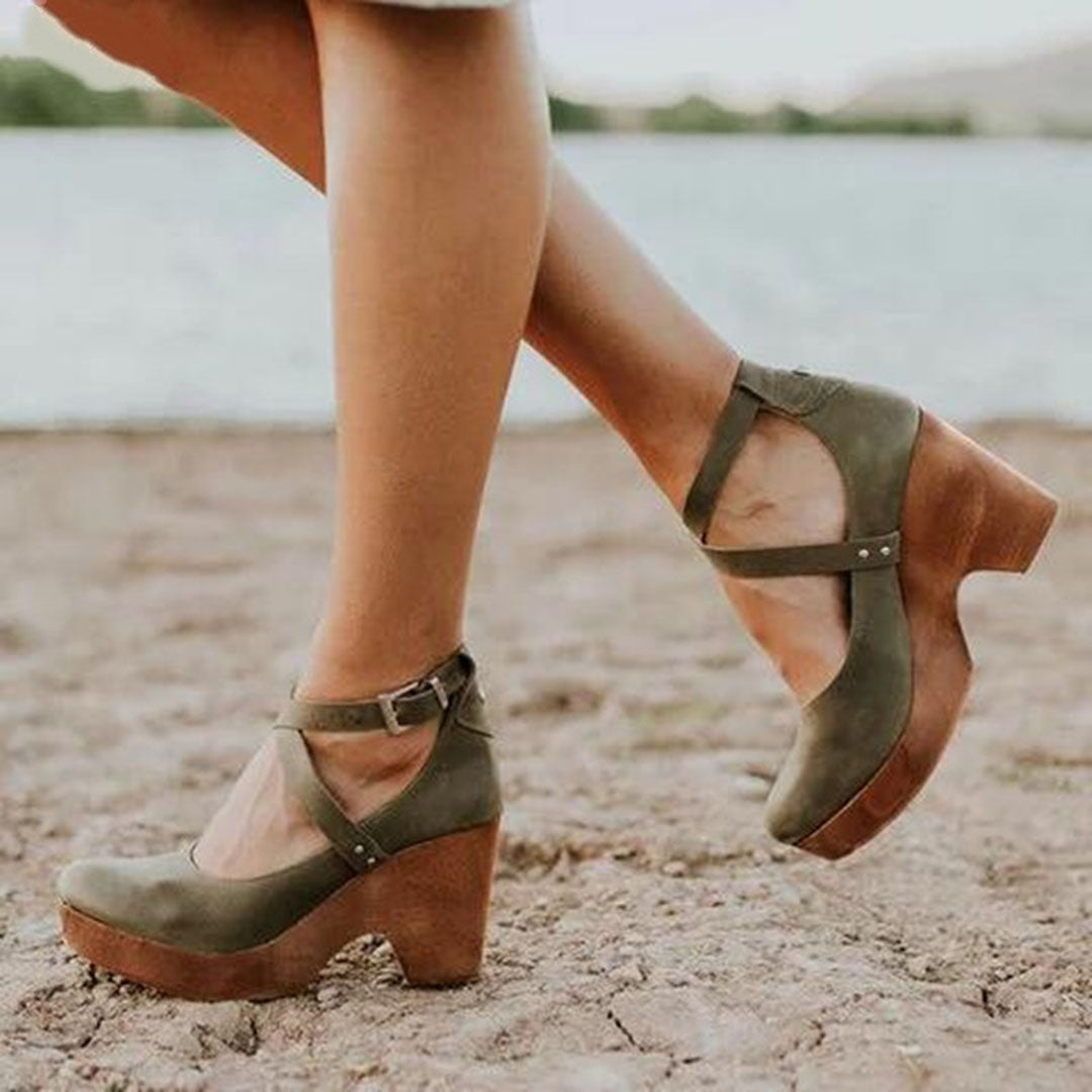 Woman showcasing ankle strap wedge shoes against a plain background.