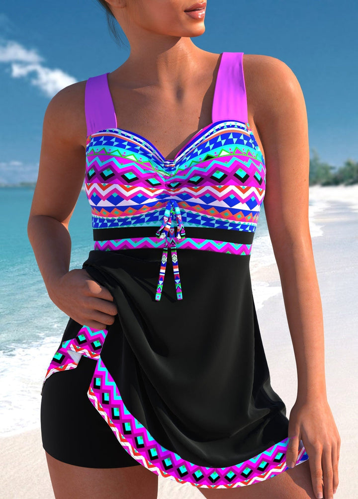 Female model wearing a colorful geometric swim dress with built-in shorts on a plain background.