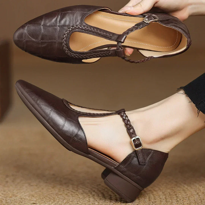 Black pointed toe Mary Jane flat with braided T-strap and buckle on a plain background.