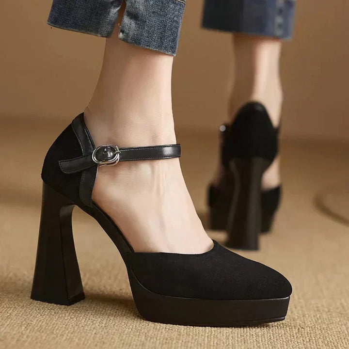 Female model wearing elevated block heel pumps with ankle straps on a plain background.