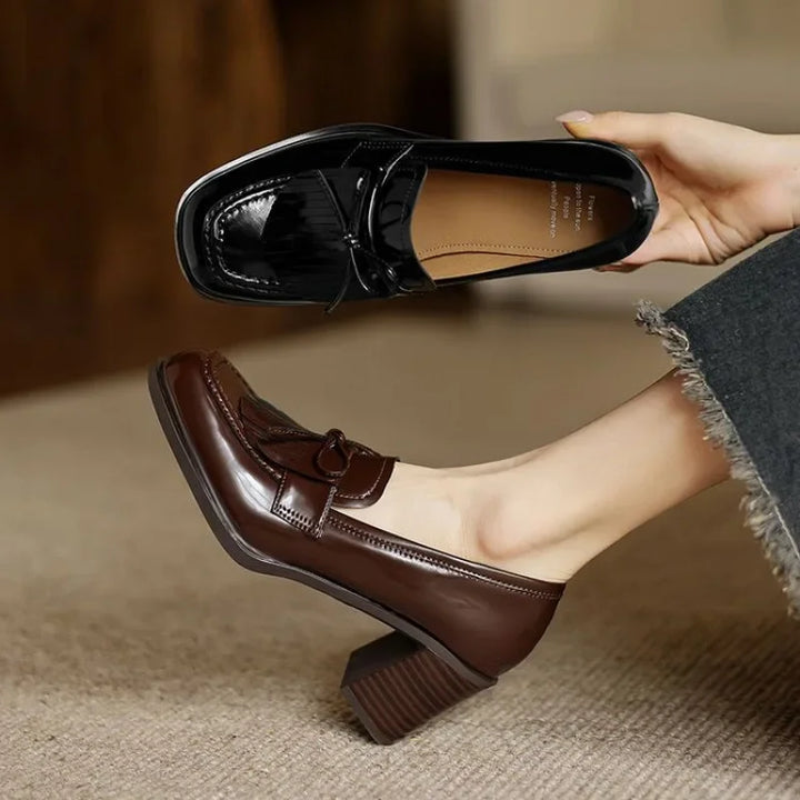 Female showcasing polished dark loafers with tassel vamp detail on a plain background.