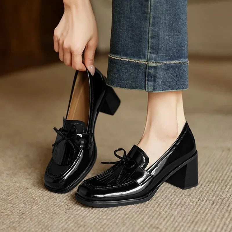 Woman wearing glossy tassel loafer pumps with a block heel on a plain background.