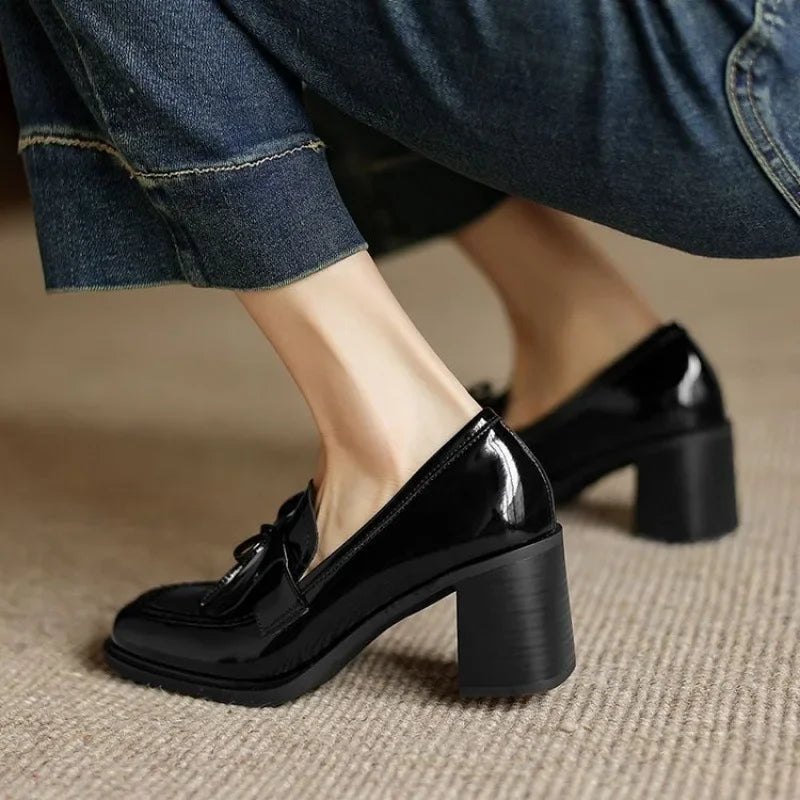 Woman showcasing polished block heel loafers with tassel detail on a plain background.