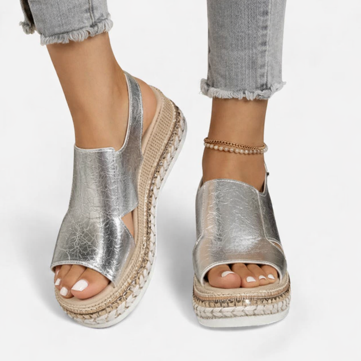 Model wearing metallic wedge sandals on a plain background with an open toe and adjustable slingback strap.