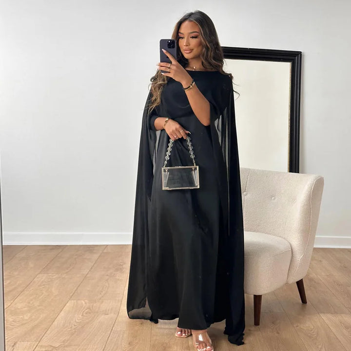 Woman in an elegant satin maxi dress with cape overlay on a plain background.