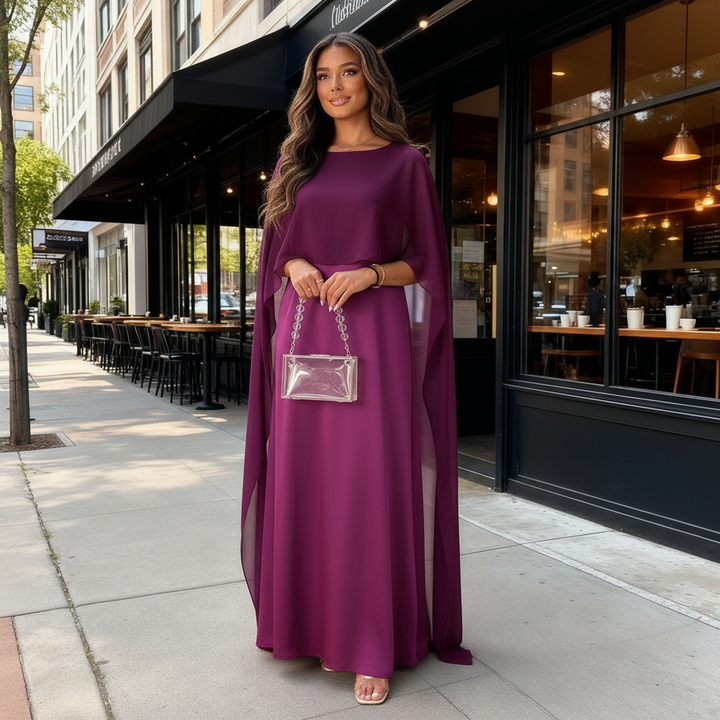 Female model in a floor-length evening dress with flowing overlay on a plain background.